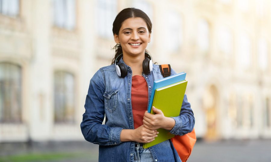 Happy pretty indian young lady student posing at university or college campus outdoors, carrying backpack and course books, smiling at camera. Education concept, copy space